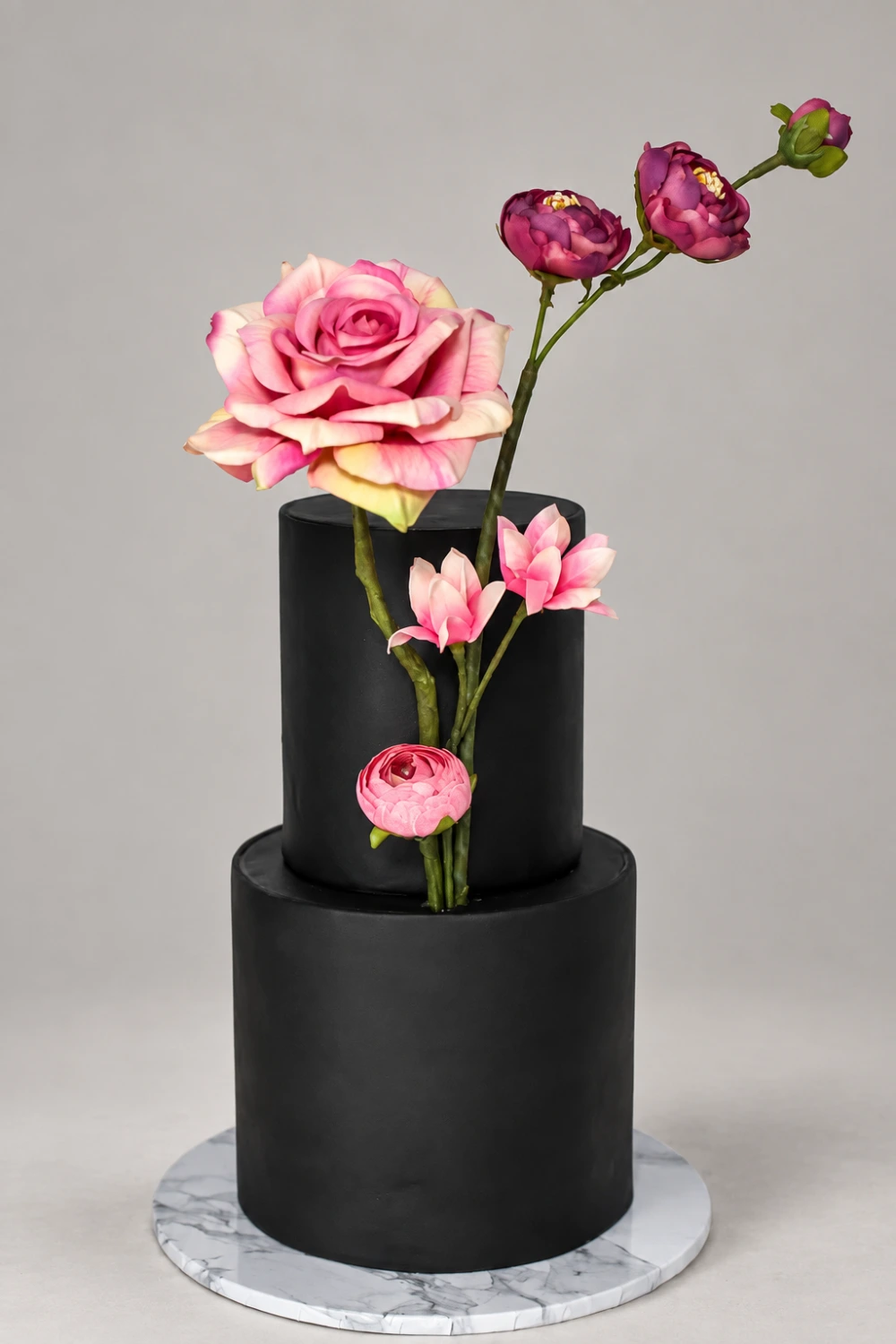 Two-tier black cake with an upright pink sugar rose and purple and pink blossoms