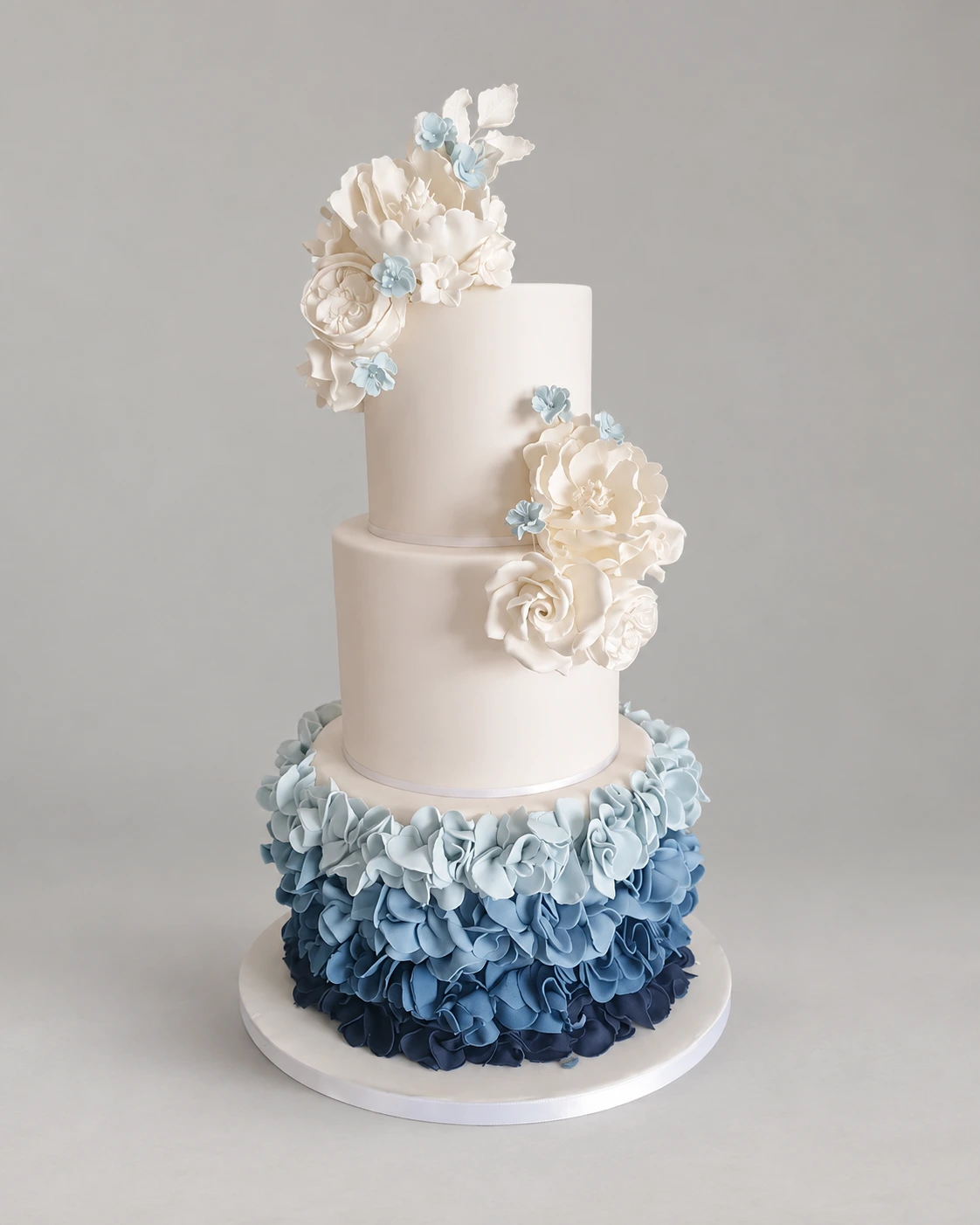 Three-tier cake with layered blue ruffles on the bottom tier and ivory sugar flower clusters