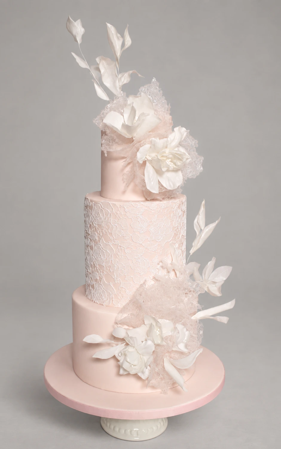 Three-tier pale blush cake with white sugar flowers, delicate lace texture and white foliage