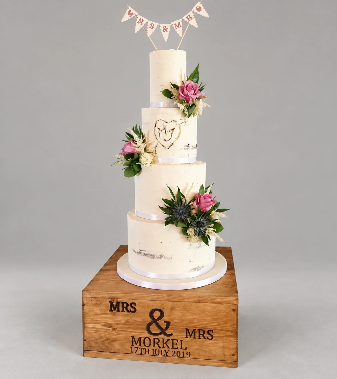 Four-tier semi-naked wedding cake with blush roses, greenery and a heart monogram