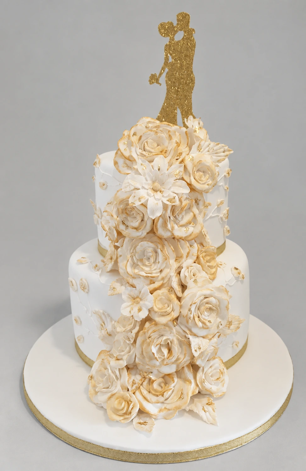Two-tier white cake covered with cascading gold-edged ivory roses and a couple topper