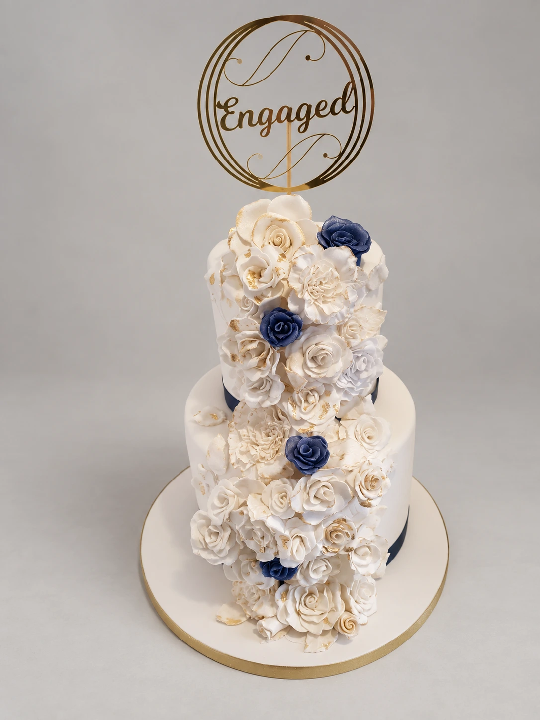 Two-tier white cake with navy ribbon, a blue and white rose cascade and an engagement topper