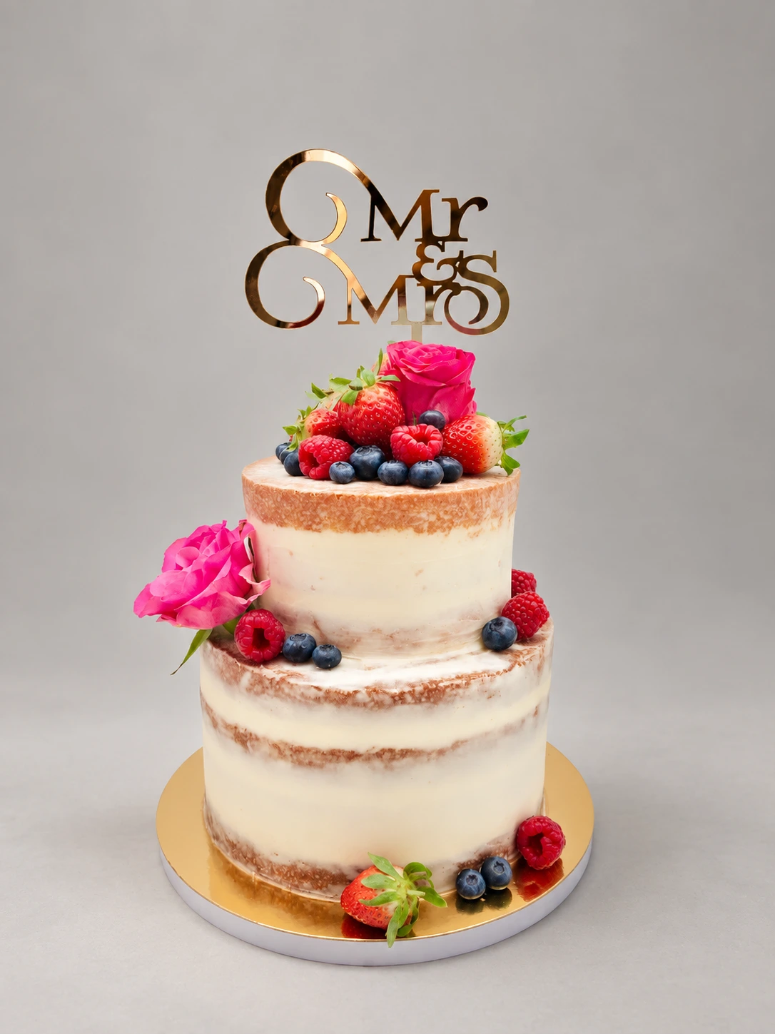 Two-tier semi-naked cake with pink fresh roses, mixed berries and a gold wedding topper
