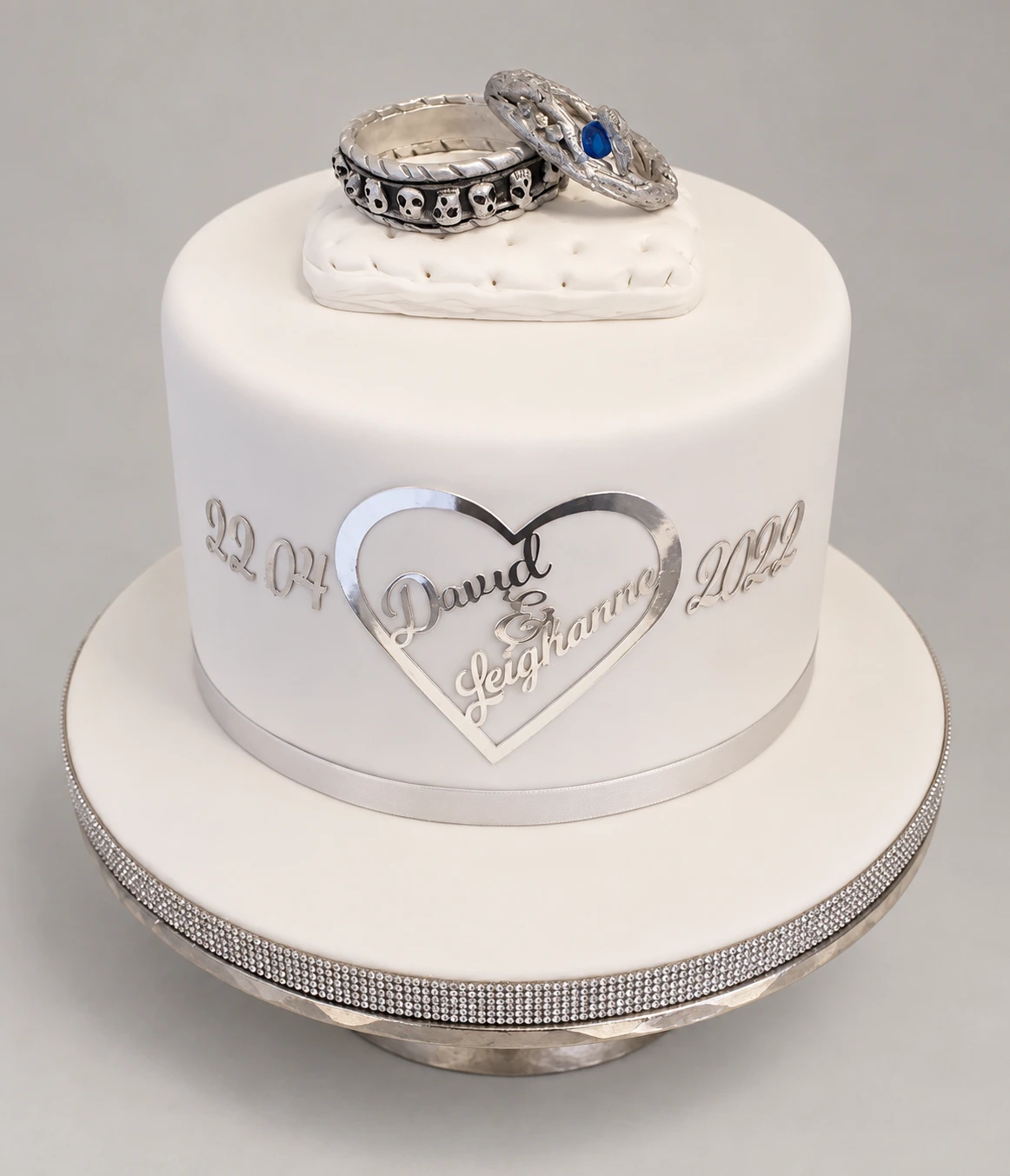Single-tier white cake with silver trim, a heart plaque and decorative rings on a small cushion