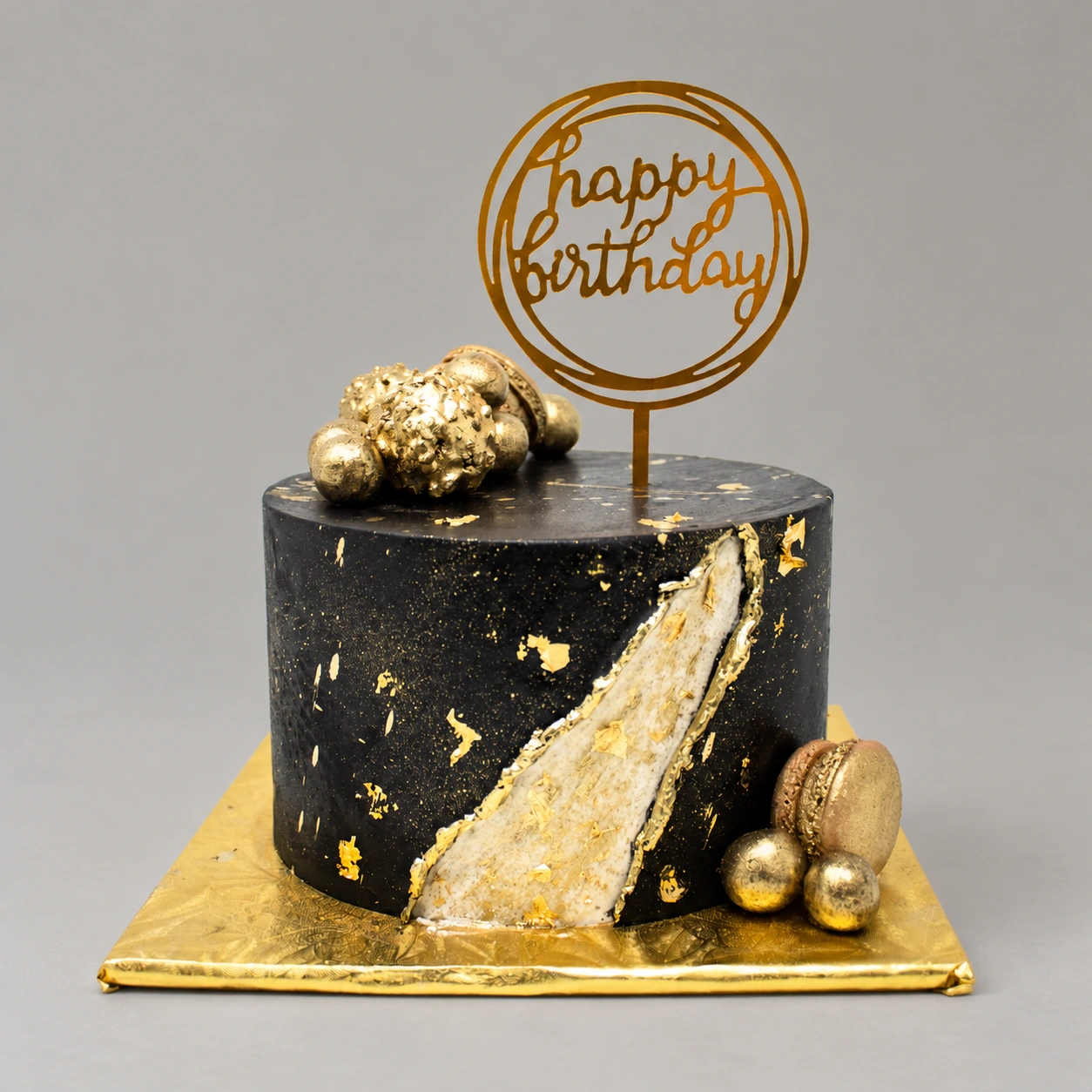 Single-tier black cake with gold leaf, an ivory and gold geode stripe, gold macarons and a birthday topper