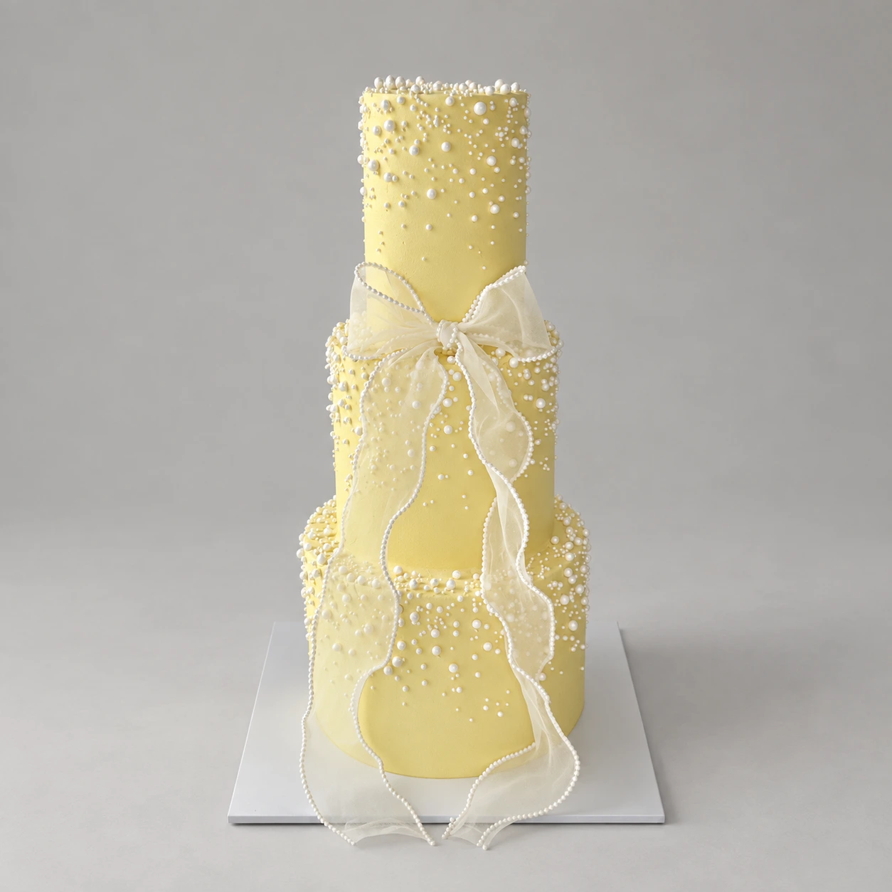 Three-tier pale yellow cake with white pearls and a long translucent ivory bow